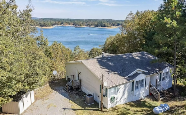 Ein eingeschossiges Haus mit geneigtem Dach und einer kleinen Veranda. Es ist von Bäumen umgeben und bietet Blick auf ein großes Gewässer im Hintergrund.
