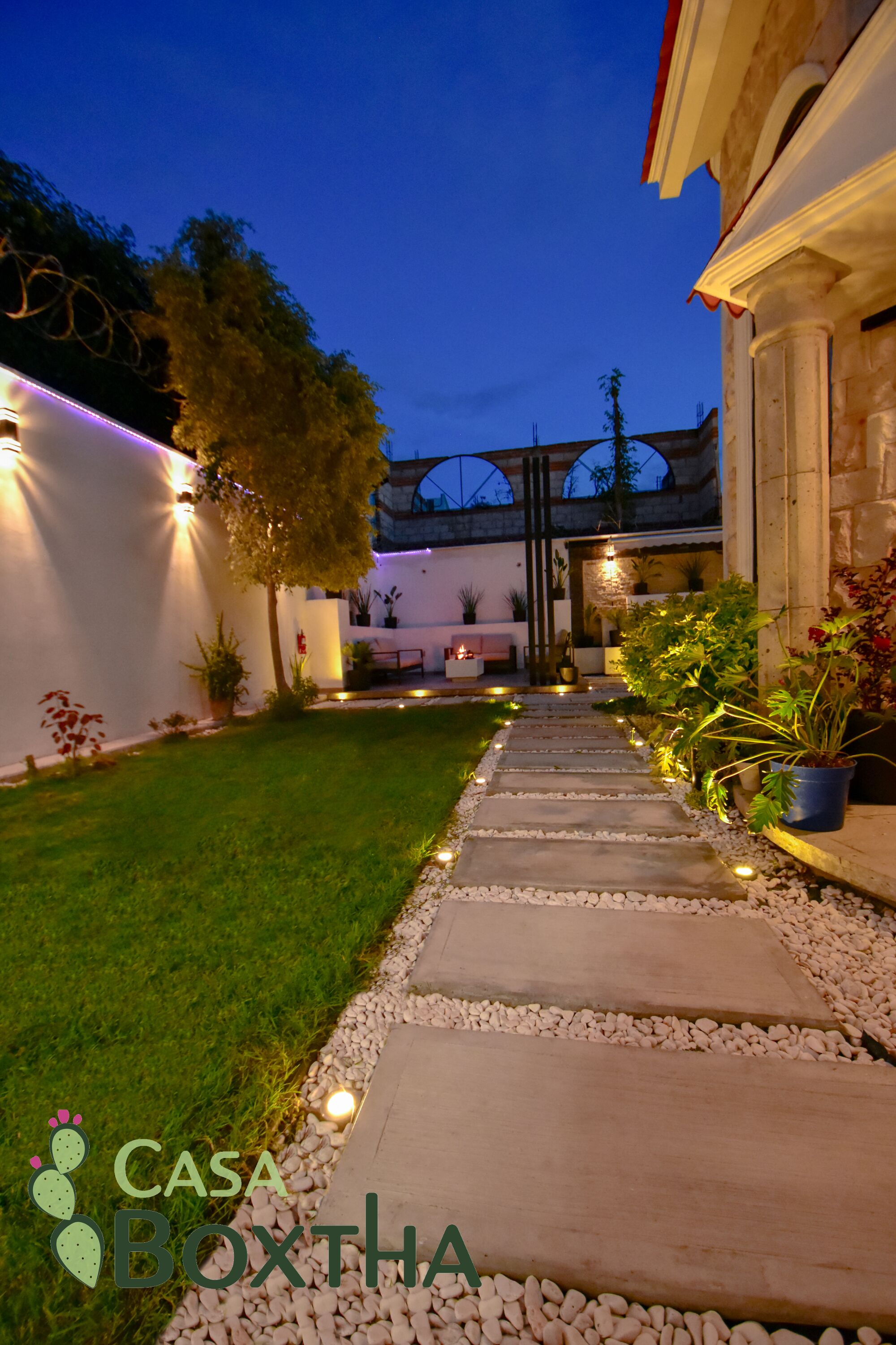 A well-lit pathway leading to a building with a garden area, potted plants, and a stone wall.