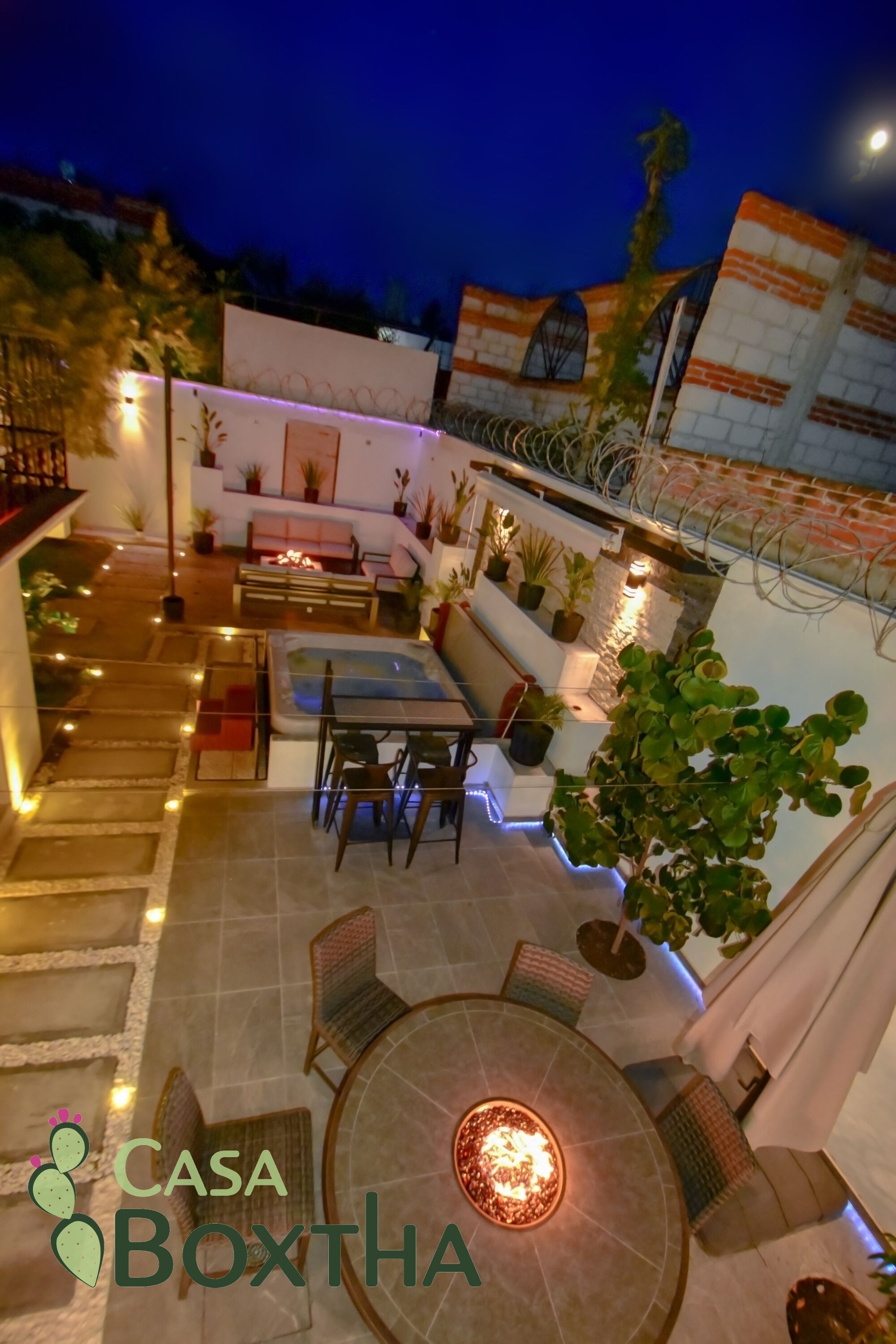 An outdoor patio area with a round table, chairs, and a sofa. There are potted plants and a fire feature.