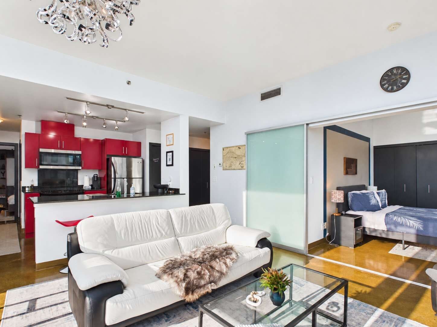 A modern living space with a white sofa, a glass coffee table, and a kitchen area featuring red cabinets and stainless steel appliances.