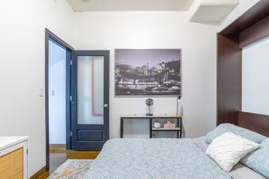 A bedroom with a bed, a nightstand, a framed cityscape picture, a door, and a small table.