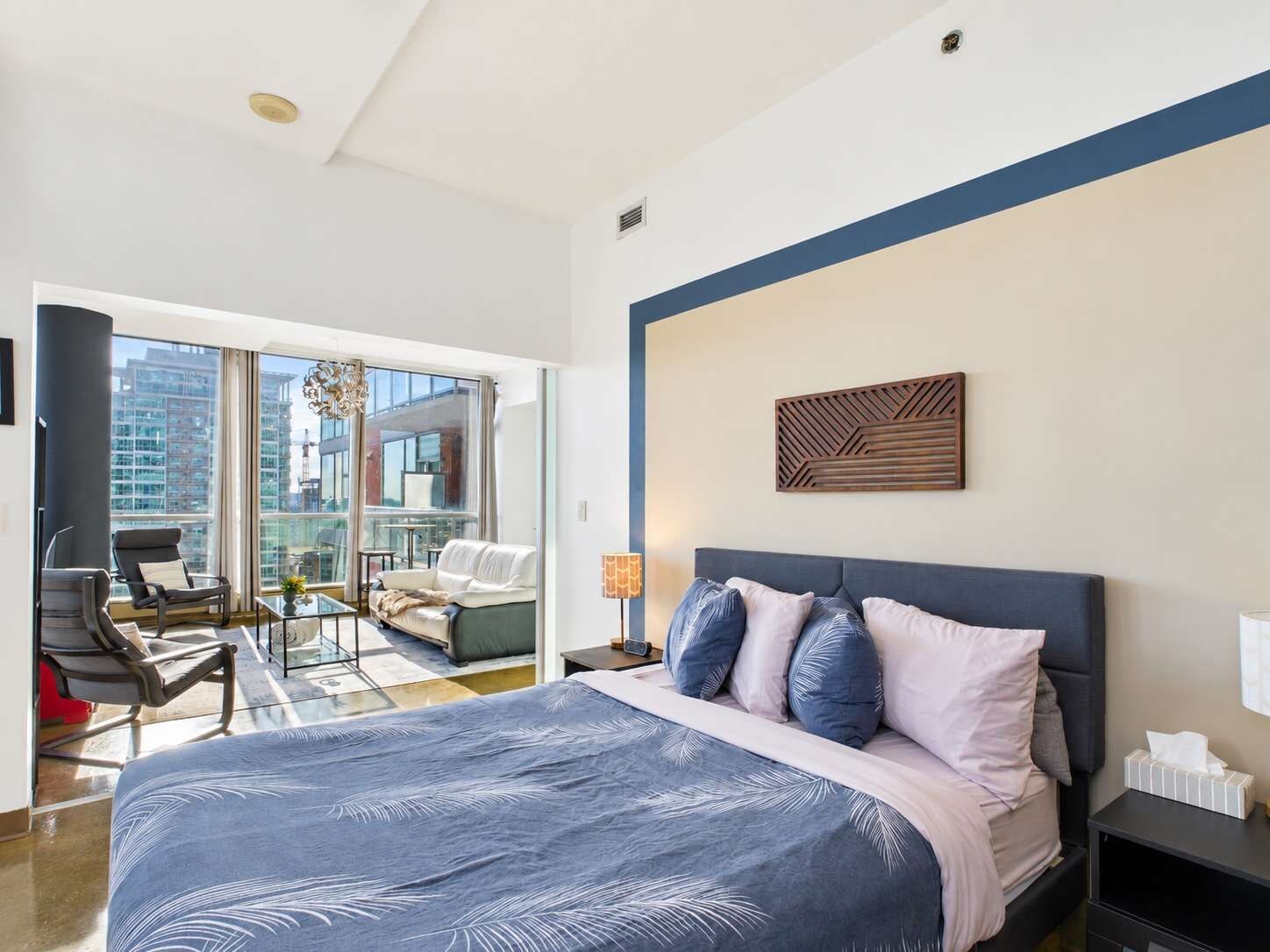 A modern bedroom with a large bed, a seating area with a sofa and chairs, and a view of the city through large windows.