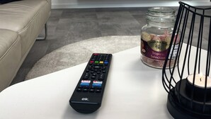 Una sala de estar con un televisor montado en la pared, un control remoto sobre una mesa blanca, un frasco de bocadillos y un candelabro.