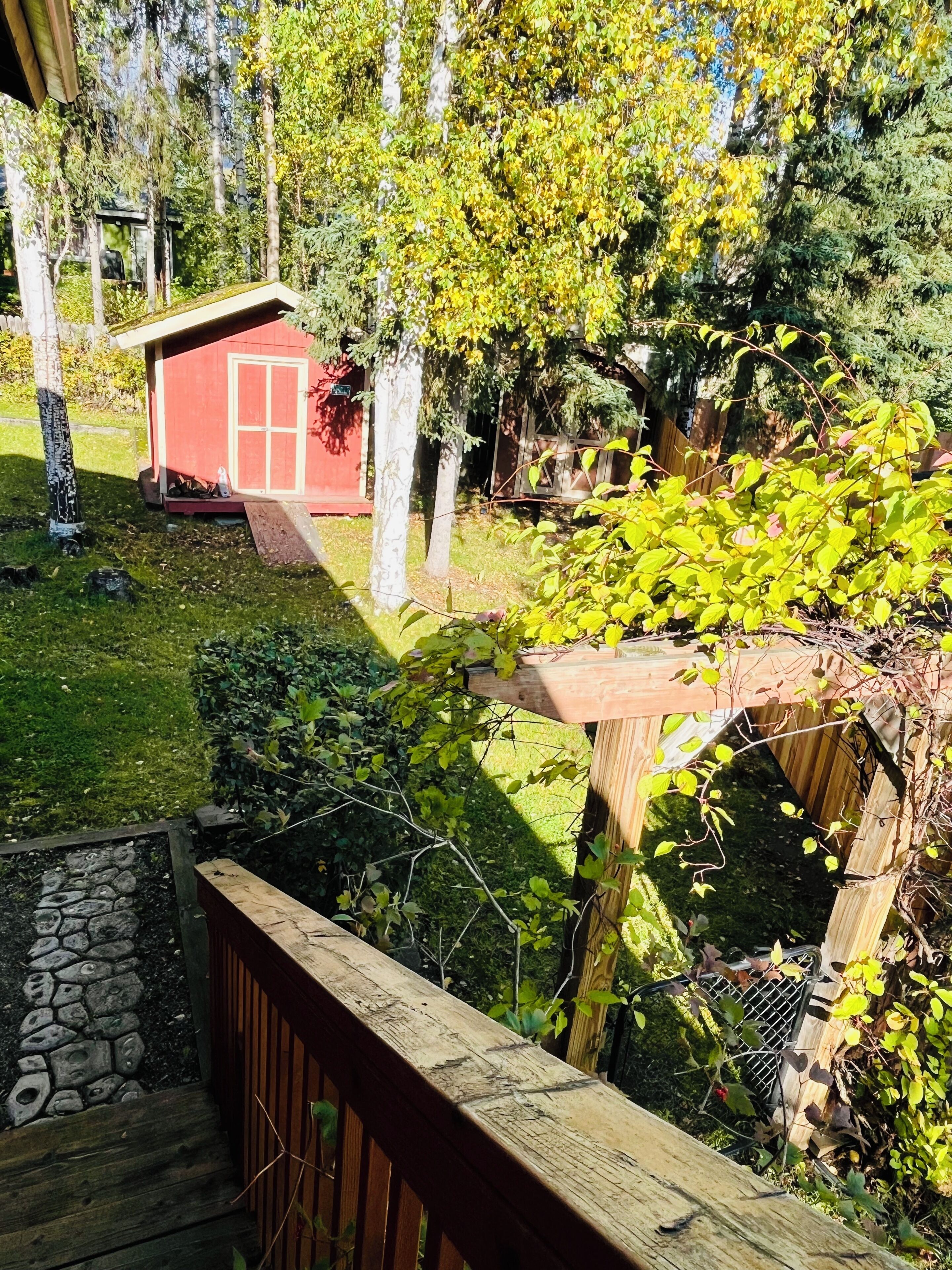 Eine Holzterrasse mit einer Steinsäule, einem roten Schuppen und einem Garten mit verschiedenen Pflanzen und Bäumen.