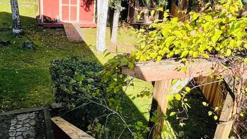 Eine Holzterrasse mit einer Steinsäule, einem roten Schuppen und einem Garten mit verschiedenen Pflanzen und Bäumen.