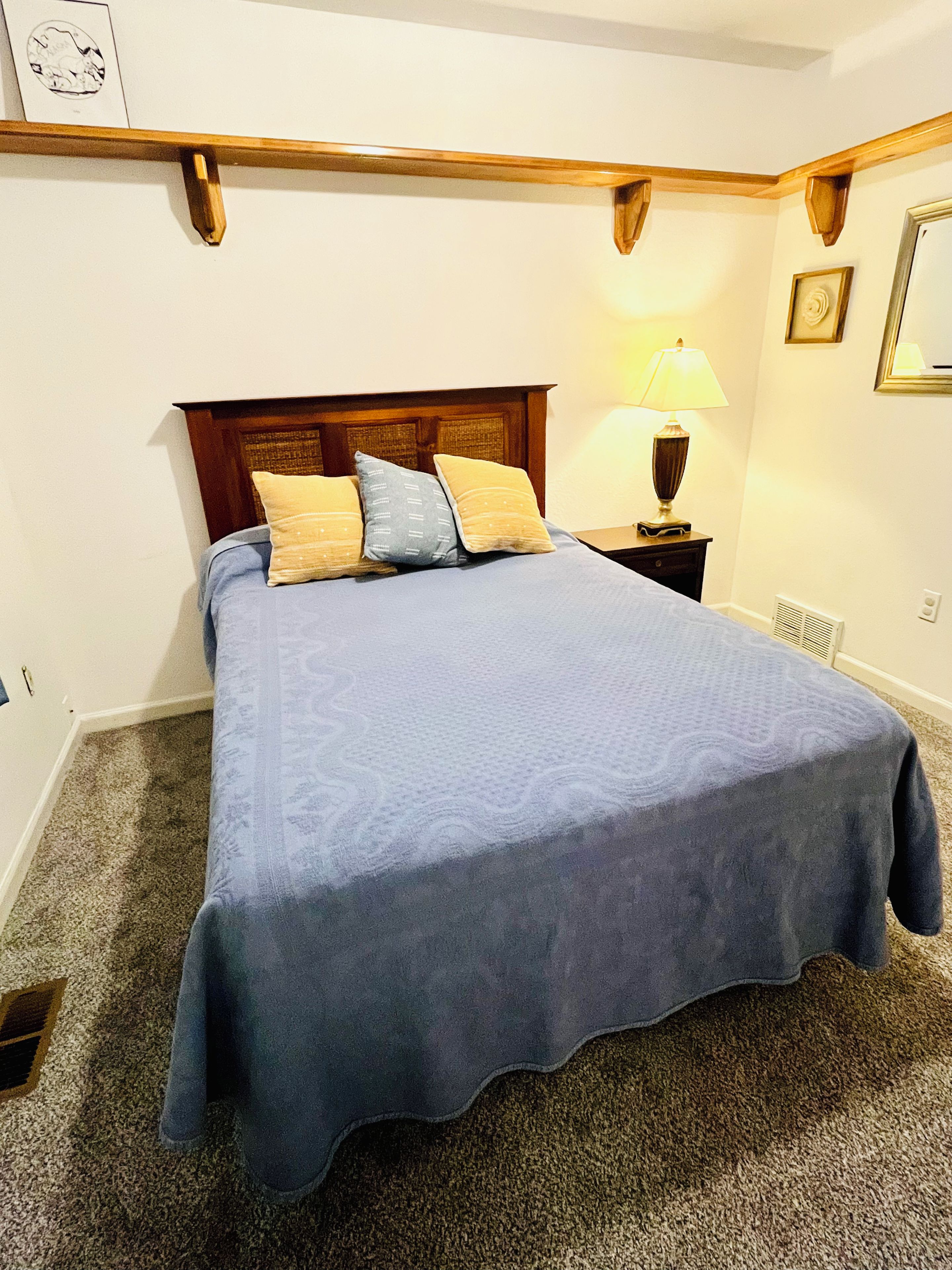 Ein ordentlich bezogenes Bett mit blauer Bettdecke und Kissen, ein Holzkopfschild, ein Nachttisch mit Lampe und ein Heizkörper auf dem Boden.