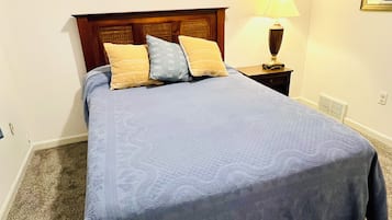 Ein ordentlich bezogenes Bett mit blauer Bettdecke und Kissen, ein Holzkopfschild, ein Nachttisch mit Lampe und ein Heizkörper auf dem Boden.