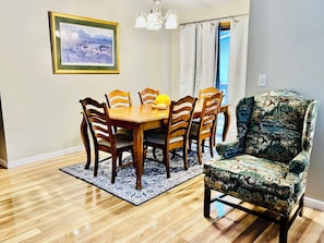 A dining area with a wooden table and chairs, a patterned rug, a framed picture on the wall, and a window with curtains.