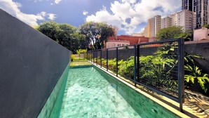 Una piscina con suelo de baldosas, rodeada por una cerca de metal negro, y vistas a edificios y árboles al fondo.
