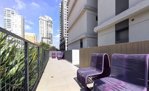 A rooftop terrace with two purple chairs, a metal fence, and tall buildings in the background. - Comfort With Balcony 100m From Puc-pompeia Puq0208 (São Paulo)