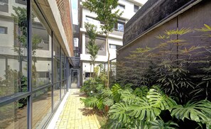 A narrow corridor with glass walls, a tiled floor, and lush tropical plants. - Studio With Balcony 100m From Puc-pompeia Puq0208 (São Paulo)