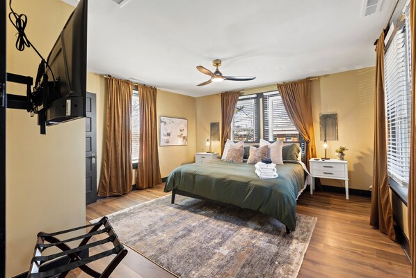 A bedroom with a bed, a ceiling fan, a TV mounted on the wall, a bedside table with a lamp, and a window with curtains.
