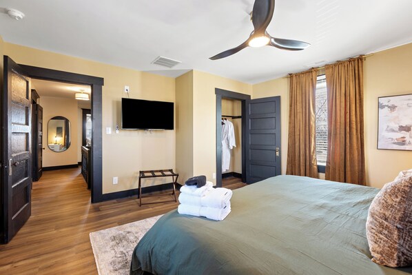 Habitación de hotel con cama, televisor de pantalla plana, ventilador de techo y vista al exterior a través de una ventana con cortinas.