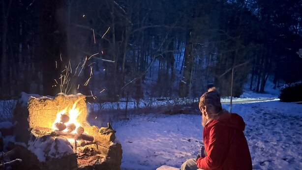 Eine Person sitzt an einem Lagerfeuer in verschneiter Winterlandschaft.