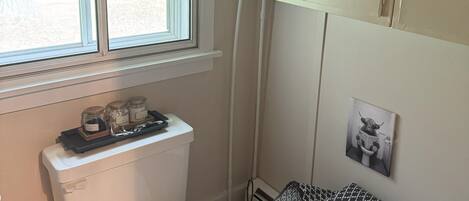 A compact bathroom with a toilet, a window, and a shelf with toiletries.