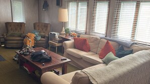 Ein Wohnzimmer mit einer Couch, einem Sessel und einem Holztisch. Es gibt Fenster mit Jalousien, eine Lampe und ein an der Wand befestigtes Regal.