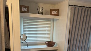 Ein Schlafzimmer mit Bett, einem Fenster mit Jalousien, einem kleinen Tisch mit Schale, einem Spiegel und einem Regal mit Bilderrahmen.