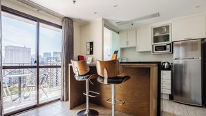 A modern kitchen with a stainless steel refrigerator, a microwave, and a wooden island. There are two bar stools with leather seats and metal frames. The room has large windows with a view of the cityscape. - Elegant Apt, Near Ibirapuera - Qsa1405 (São Paulo)