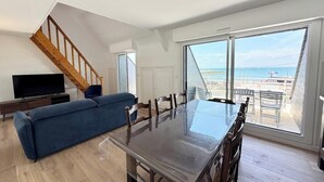 A spacious living area with a large dining table, a blue sofa, and a TV. A staircase leads to an upper level. - Duplex plein charme – Face mer & déco moderne (Quiberon)