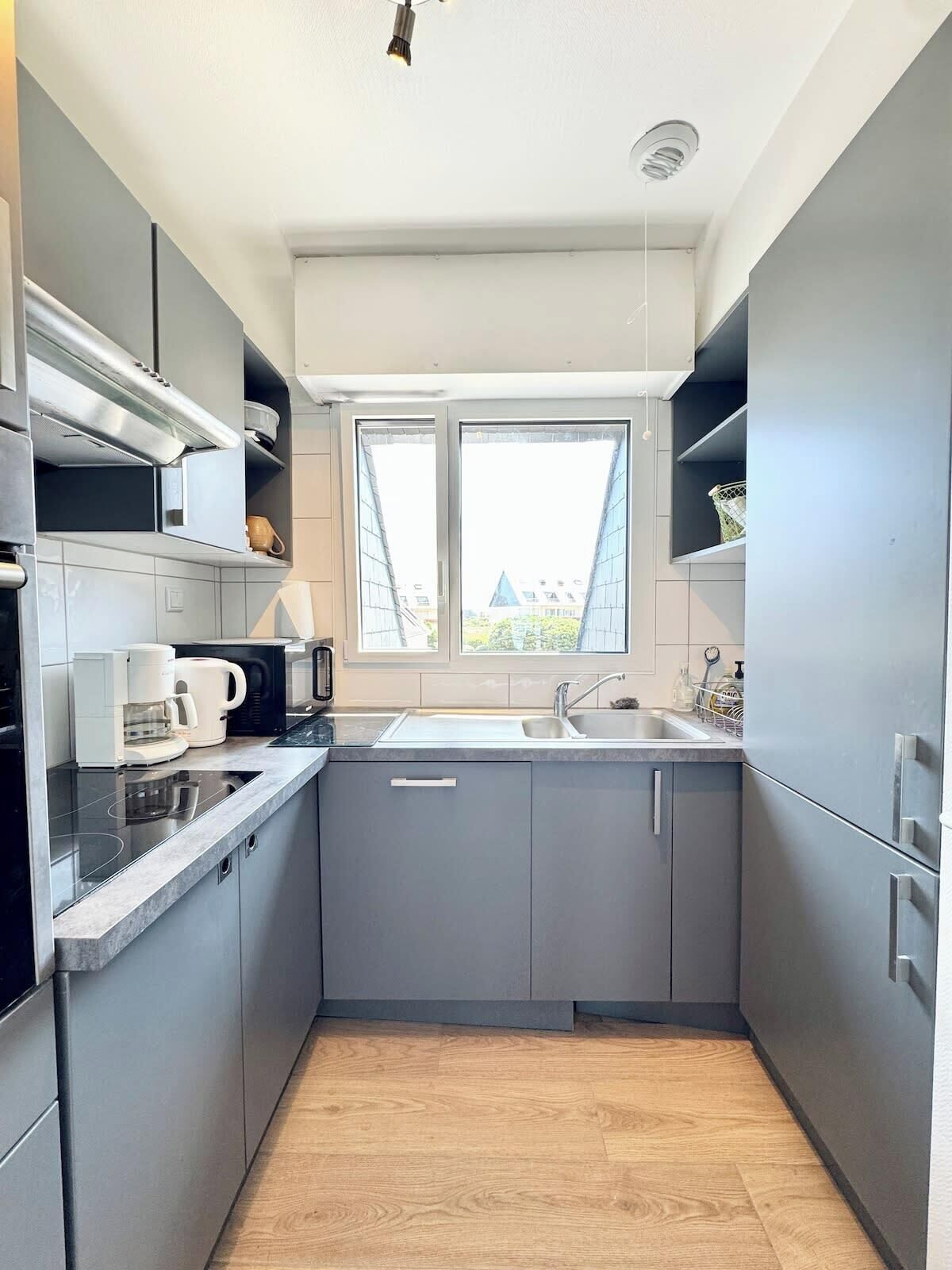 Eine moderne Küche mit grauen Schränken, einem eingebauten Backofen, einer Mikrowelle, einer Kaffeemaschine und einem Fenster mit Blick auf Gebäude.