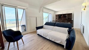 A spacious living room with a large sofa, a chair, and a kitchen area visible through an open door. - Duplex plein charme – Face mer & déco moderne (Quiberon)