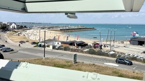 A coastal view from a window overlooking a beach, a parking area, and a marina with boats. - Duplex plein charme – Face mer & déco moderne (Quiberon)