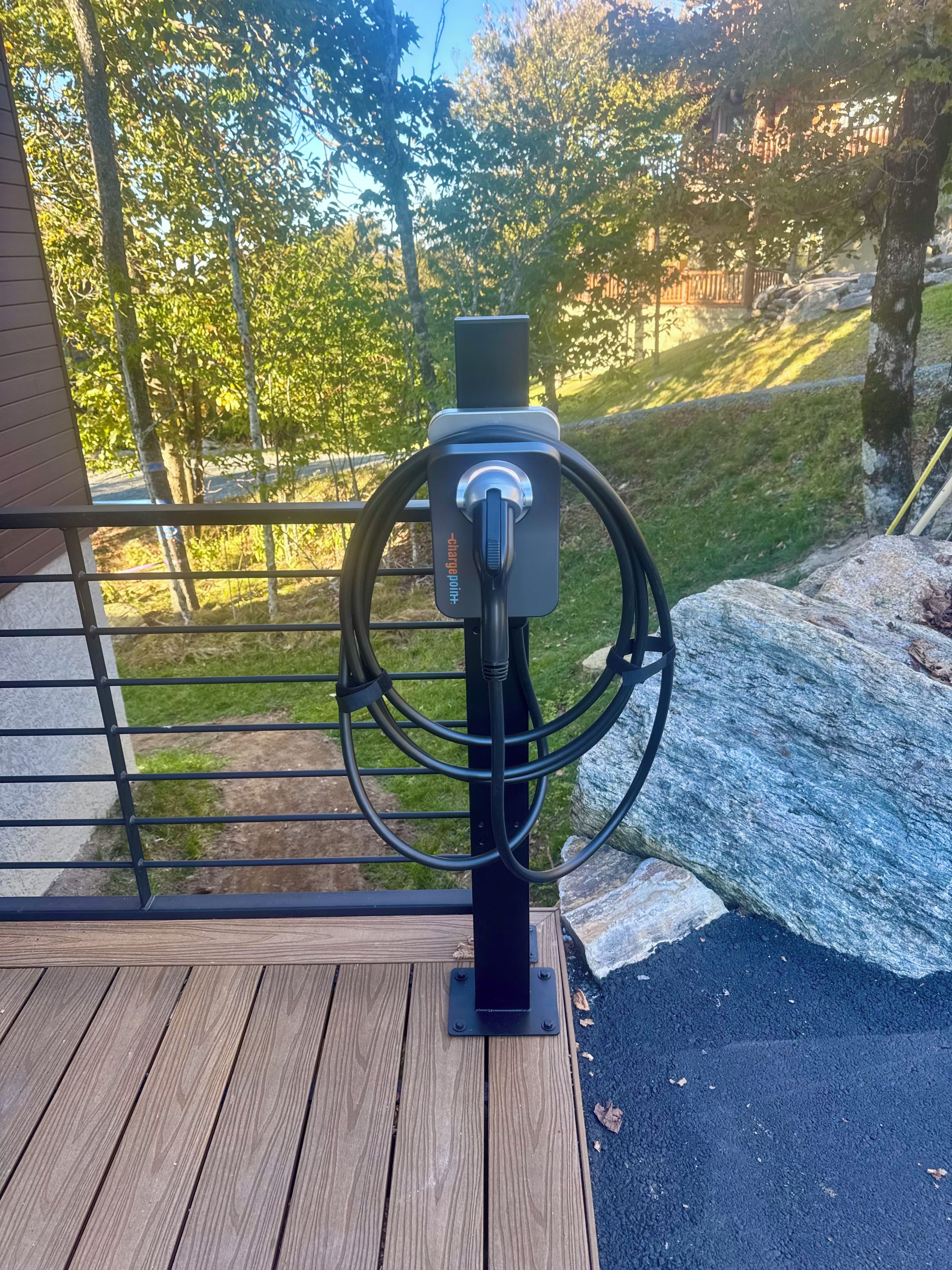 A power outlet with a plug on a wooden deck with a metal railing, overlooking a grassy area with trees.