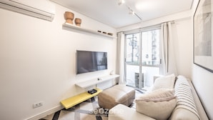 A modern living room with a flat-screen TV, a sofa, a coffee table, and a window with a city view. - Functional Apartment in Itaim Bibi SP - Sat0012 (São Paulo)