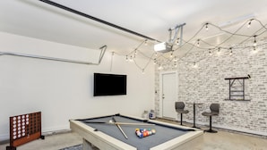 A pool table with balls, a TV, and string lights in a room with brick-patterned walls and a door.