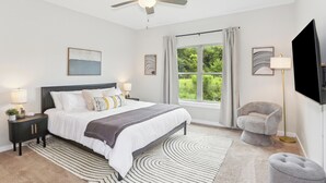 A bedroom with a bed, a ceiling fan, a window with curtains, a TV, and a side table with a lamp.