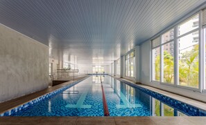Une piscine intérieure avec des carreaux bleu clair, de grandes fenêtres et une vue sur les palmiers à l’extérieur.