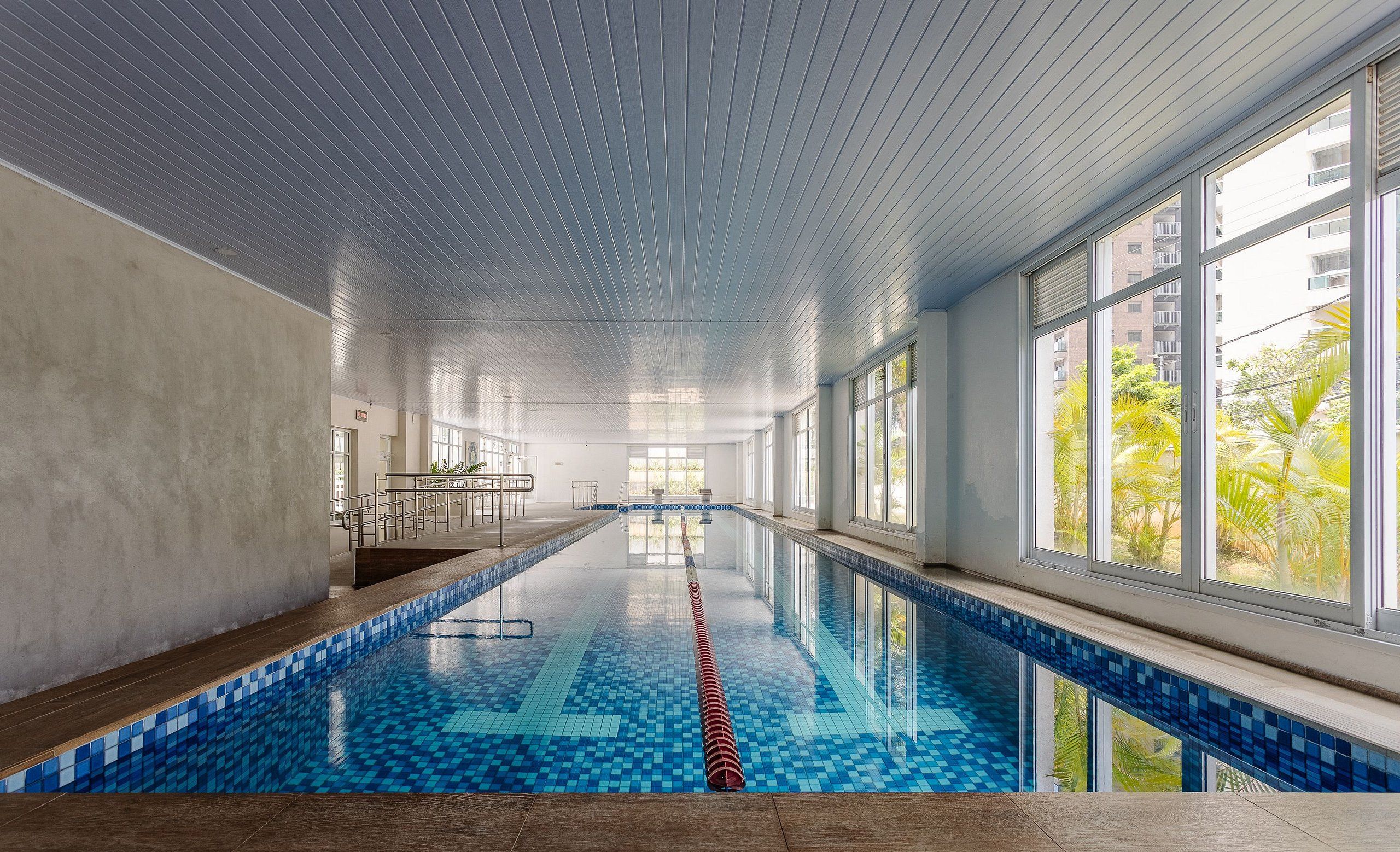 An indoor swimming pool with clear blue tiles, large windows, and a view of palm trees outside.