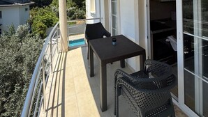 Een balkon met een tafel en twee stoelen, een schuifdeur en uitzicht op een zwembad en omliggende huizen.