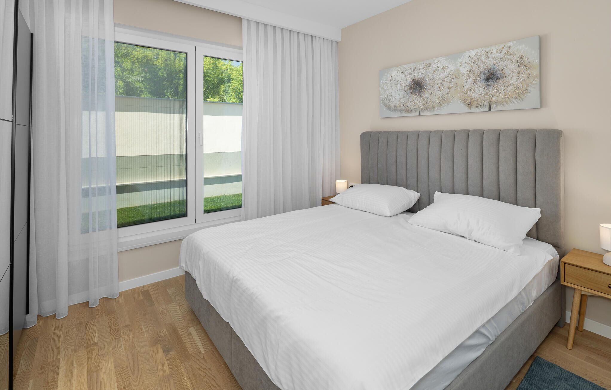 A bedroom with a large bed, white bedding, a grey headboard, a window with sheer curtains, and a wall-mounted abstract art piece.