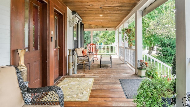 A covered porch with wooden flooring, a white railing, and outdoor furniture including a wicker chair and a table. - Lumberman's Suite at Newaygo Bed & Breakfast (Newaygo)