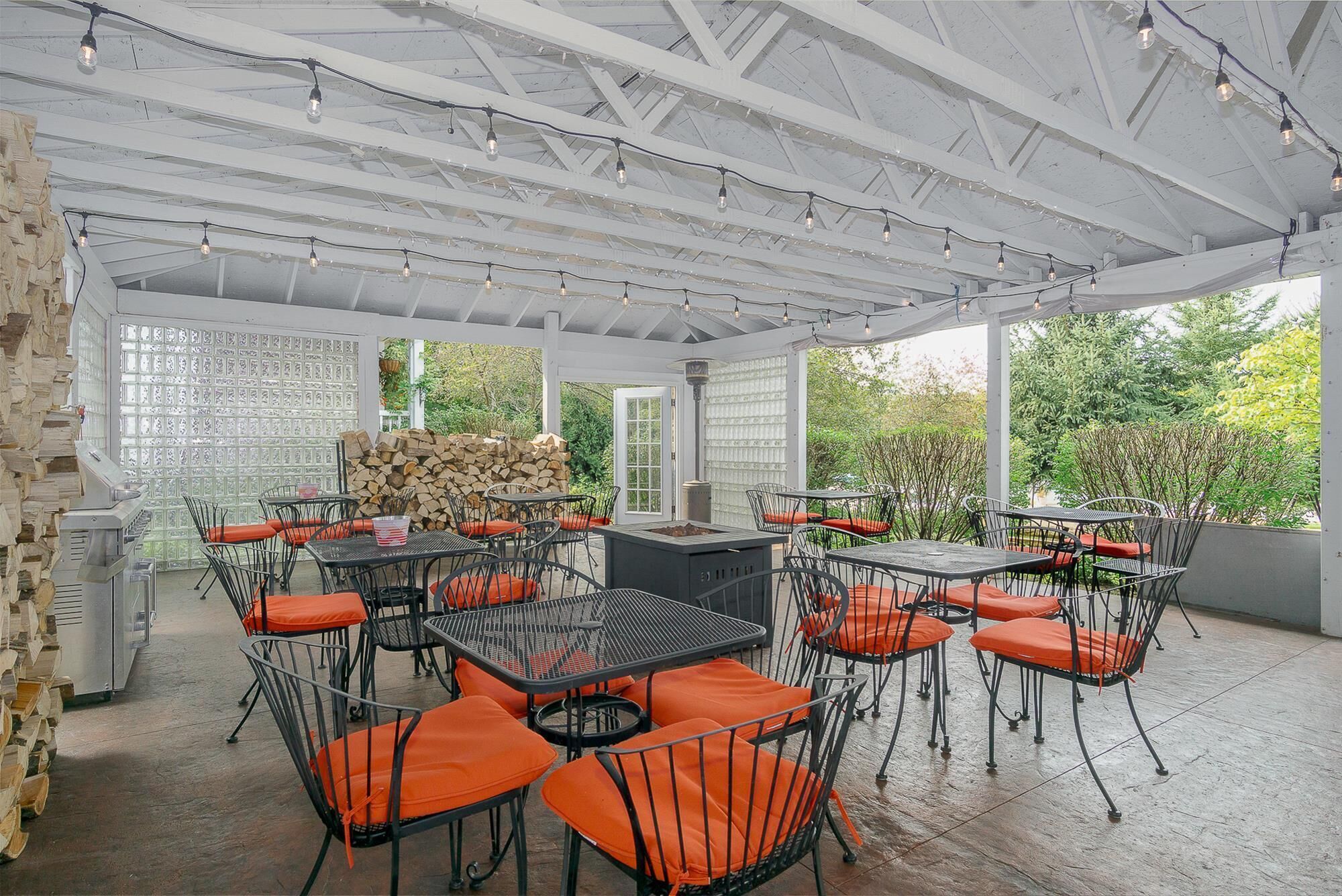 Une terrasse extérieure avec des tables et des chaises en métal, un barbecue et une pile de bois de chauffage.