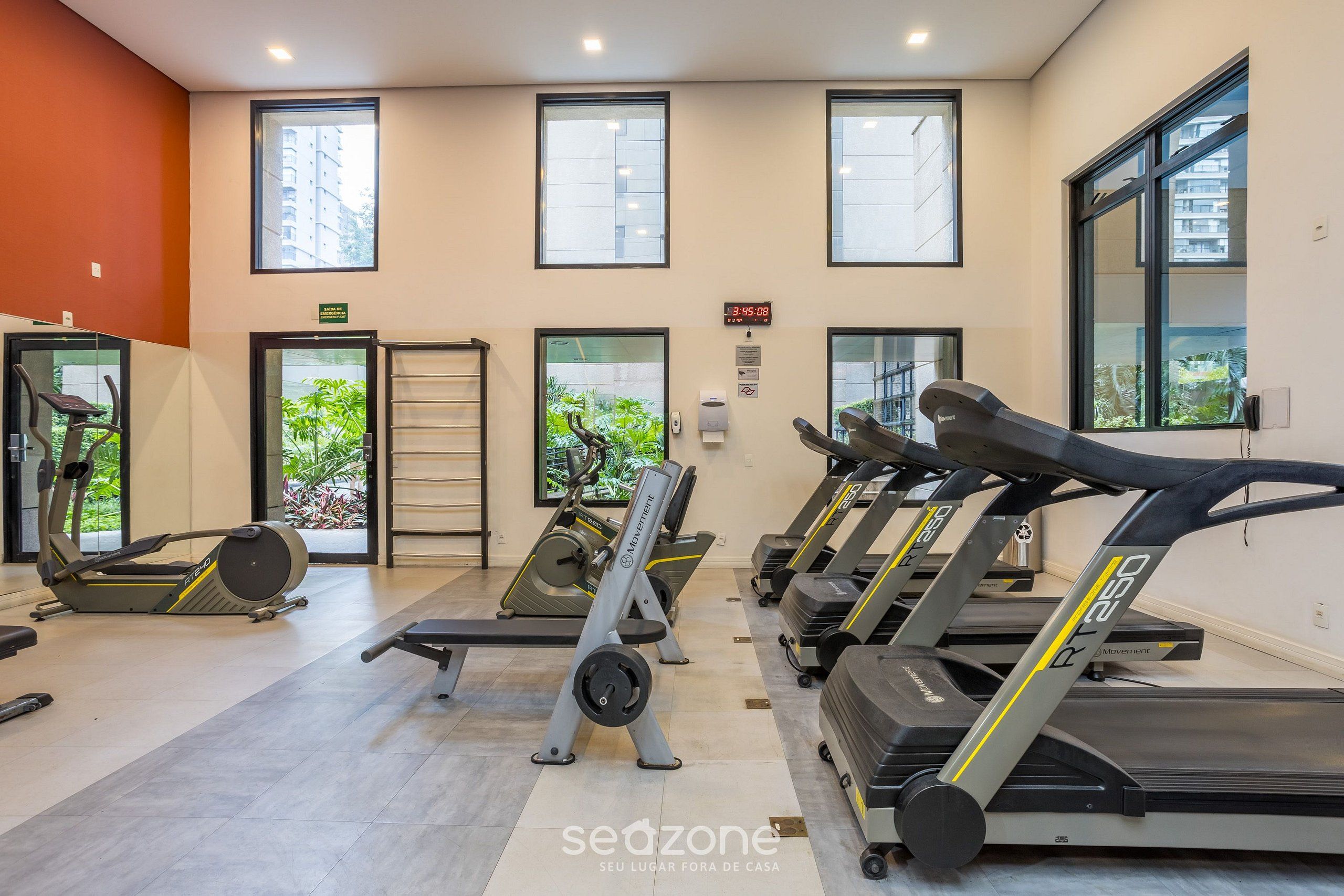Un gimnasio con cintas de correr, elípticas y bancos de pesas. Hay ventanas amplias y se ve follaje verde al exterior.