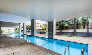 Una piscina con agua azul cristalina, rodeada de sillas de descanso y sombrillas, con suelo de hormigón y un muro bajo con plantas.