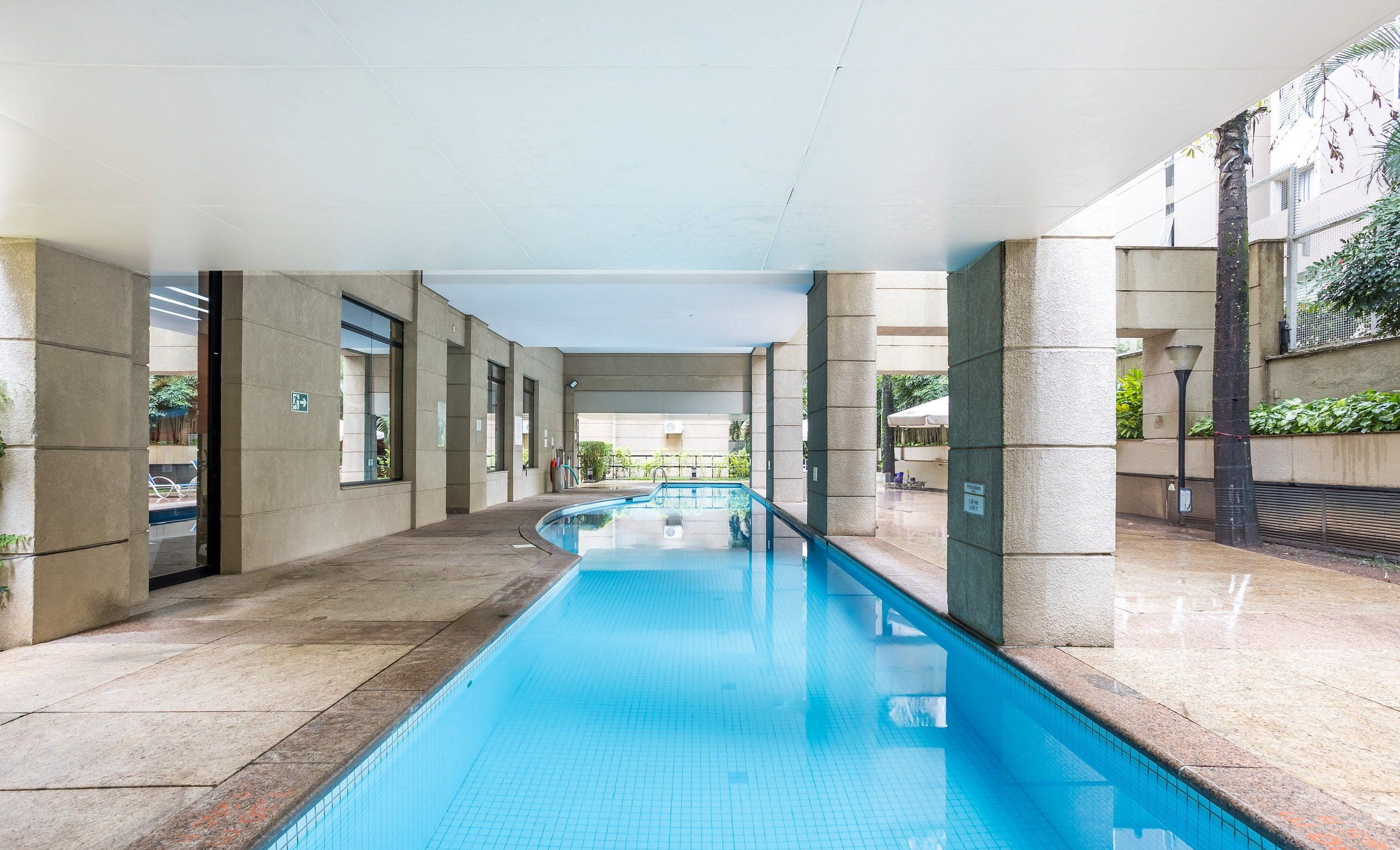 Uma piscina com água azul cristalina, rodeada por uma galeria coberta com pilares de pedra e portas de vidro.