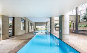 Una piscina con agua azul cristalina, rodeada por un paseo techado con pilares de piedra y puertas de cristal.
