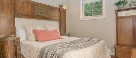 A bedroom with a large bed, wooden headboard, nightstands, and a window with a view of greenery.