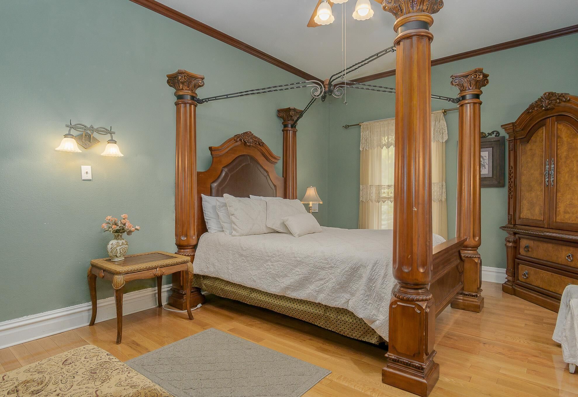 Una cama con dosel, un cabecero de madera, una mesita de noche con un jarrón de flores y un aparador de madera.