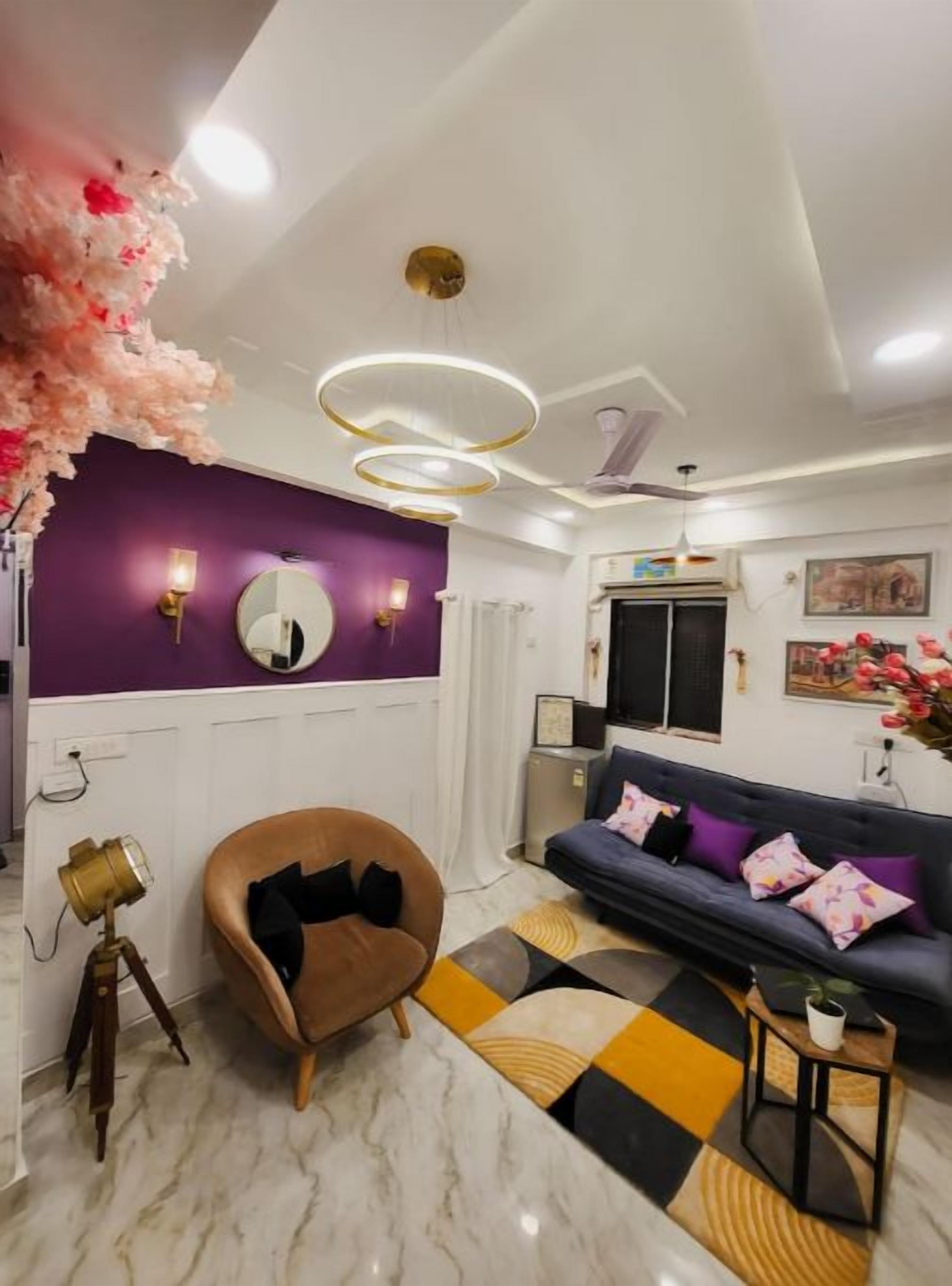 A modern living room with a purple accent wall, a brown armchair, a sofa with colorful cushions, and a patterned rug.