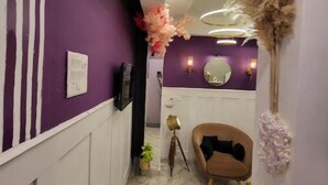 A hallway with a purple wall, a white door, a mirror, a round table, a chair, and a plant. - Amour Bandra: A Cozy Affair at Chapel Rd (Mumbai)