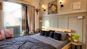 A bedroom with a large bed, a ceiling fan, a window with curtains, and a small table with a plant. - Amour Bandra: A Cozy Affair at Chapel Rd (Mumbai)