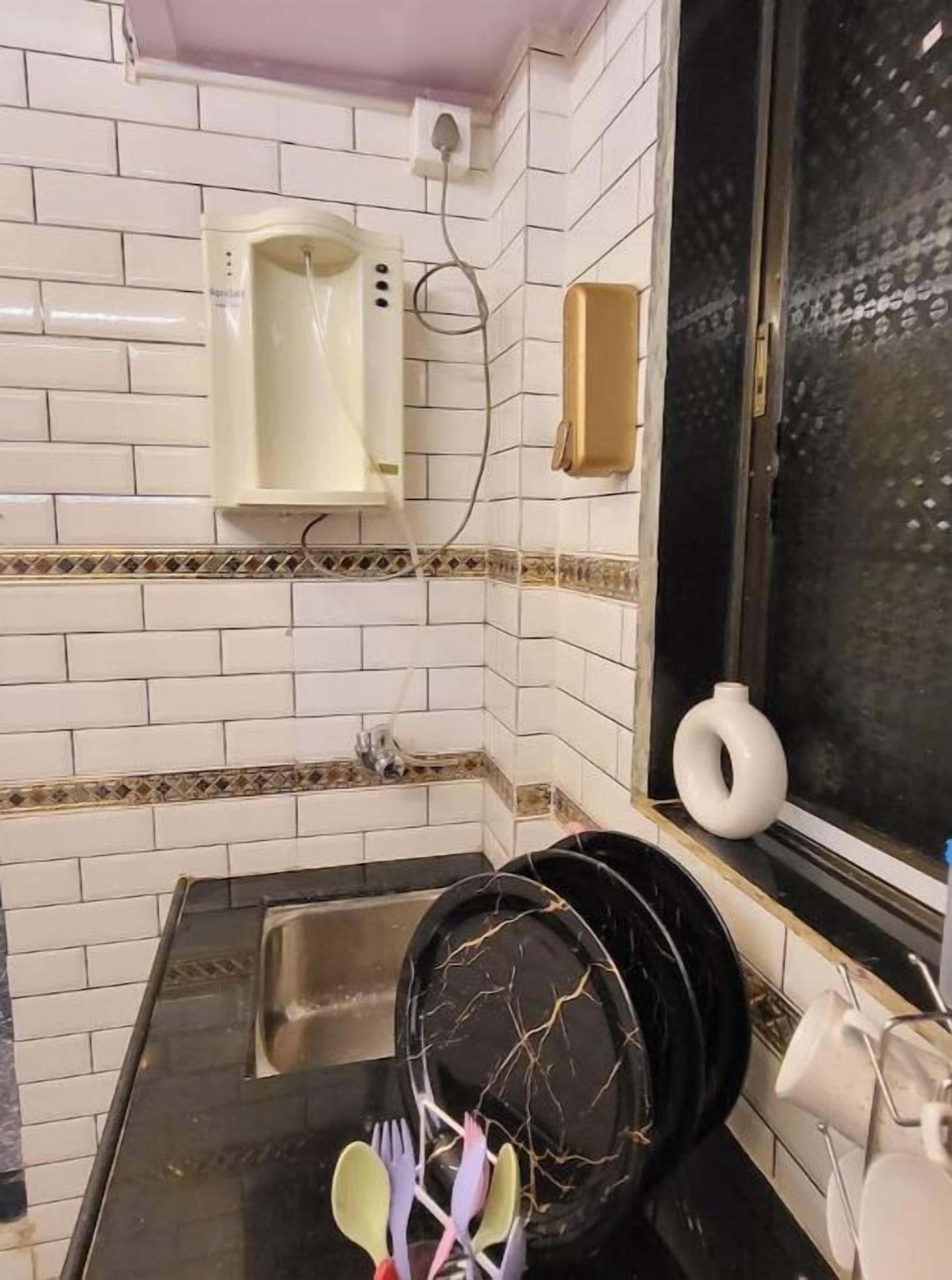 Una cocina con paredes de azulejos blancos, una encimera negra, un lavabo y una ventana con un jarrón blanco decorativo.