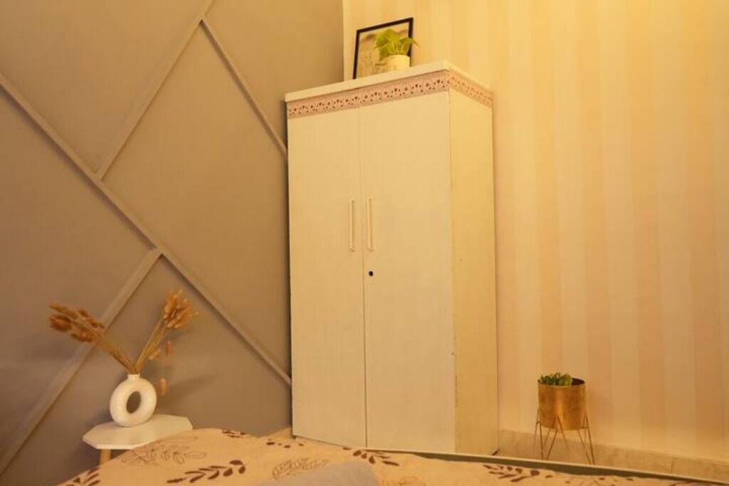 A room with a white wardrobe, a small table with a vase, and a plant on a stand.