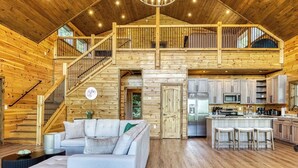 A spacious log cabin interior with a living area, kitchen, and a staircase leading to a loft.
