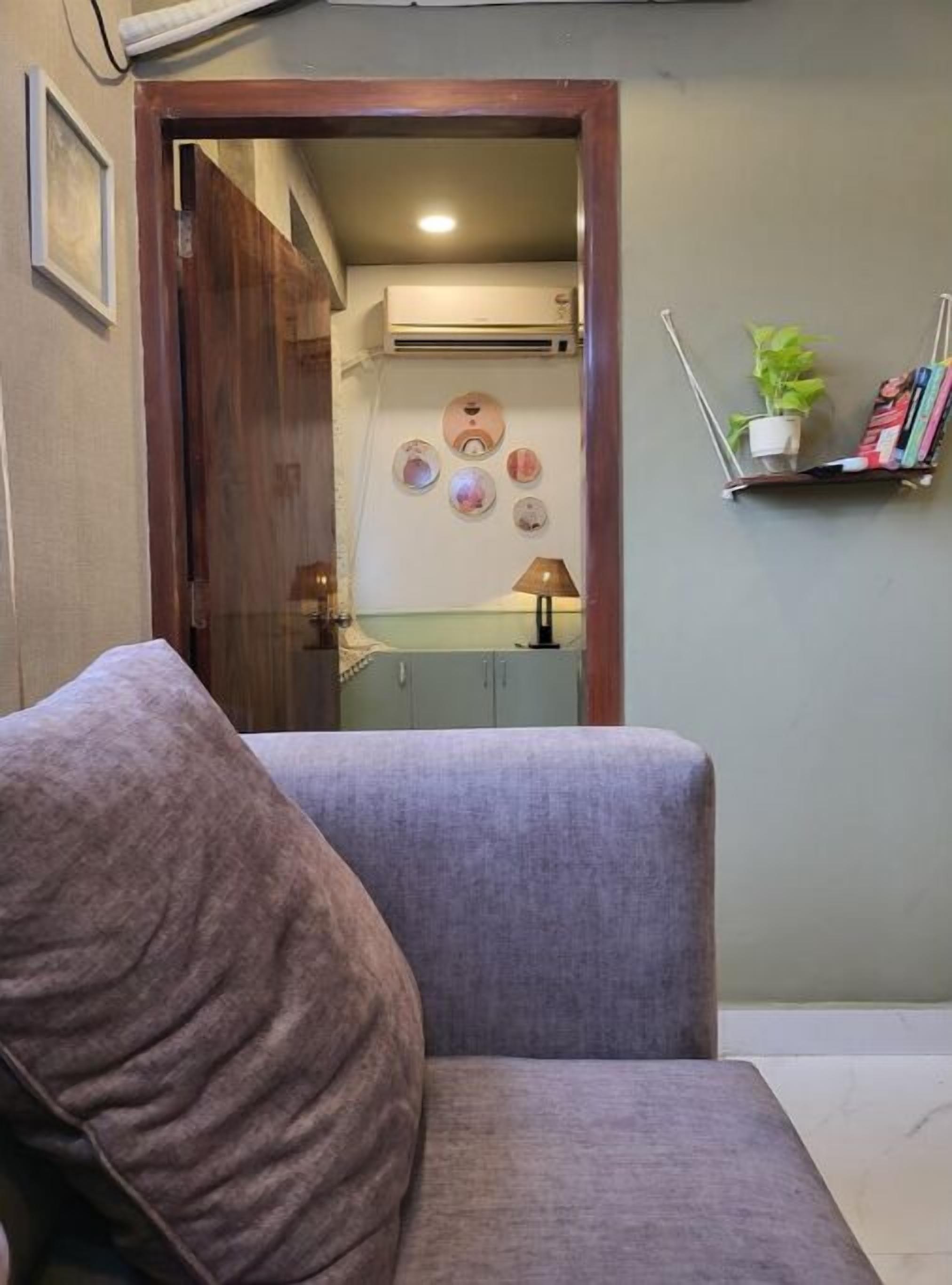 A living room with a grey sofa, a wooden door leading to another room, a wall-mounted shelf with books and a plant, and a wall with decorative plates.