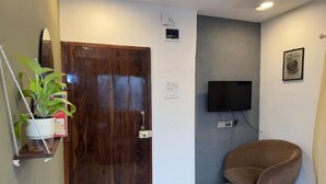 Una habitación con puerta de madera, televisor montado, una silla marrón y una planta en maceta.